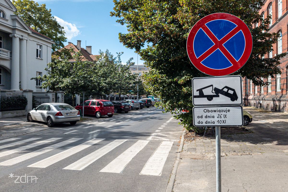 Znak zakazu zatrzymywania się z tablczką o odholowaniu pojazdów. W tle drzewa i parkujące samochody