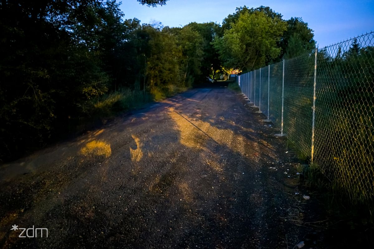 zdjęcie wykonane po ciemku, po lewej zabudowania i zaparkowane pojazdy wzdłuż płotu, centralnie ulica bez oświetlenia, po prawej płot