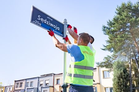 Dwaj pracownicy montują tabliczkę z nazwą ulicy Sędziwoja na słupku