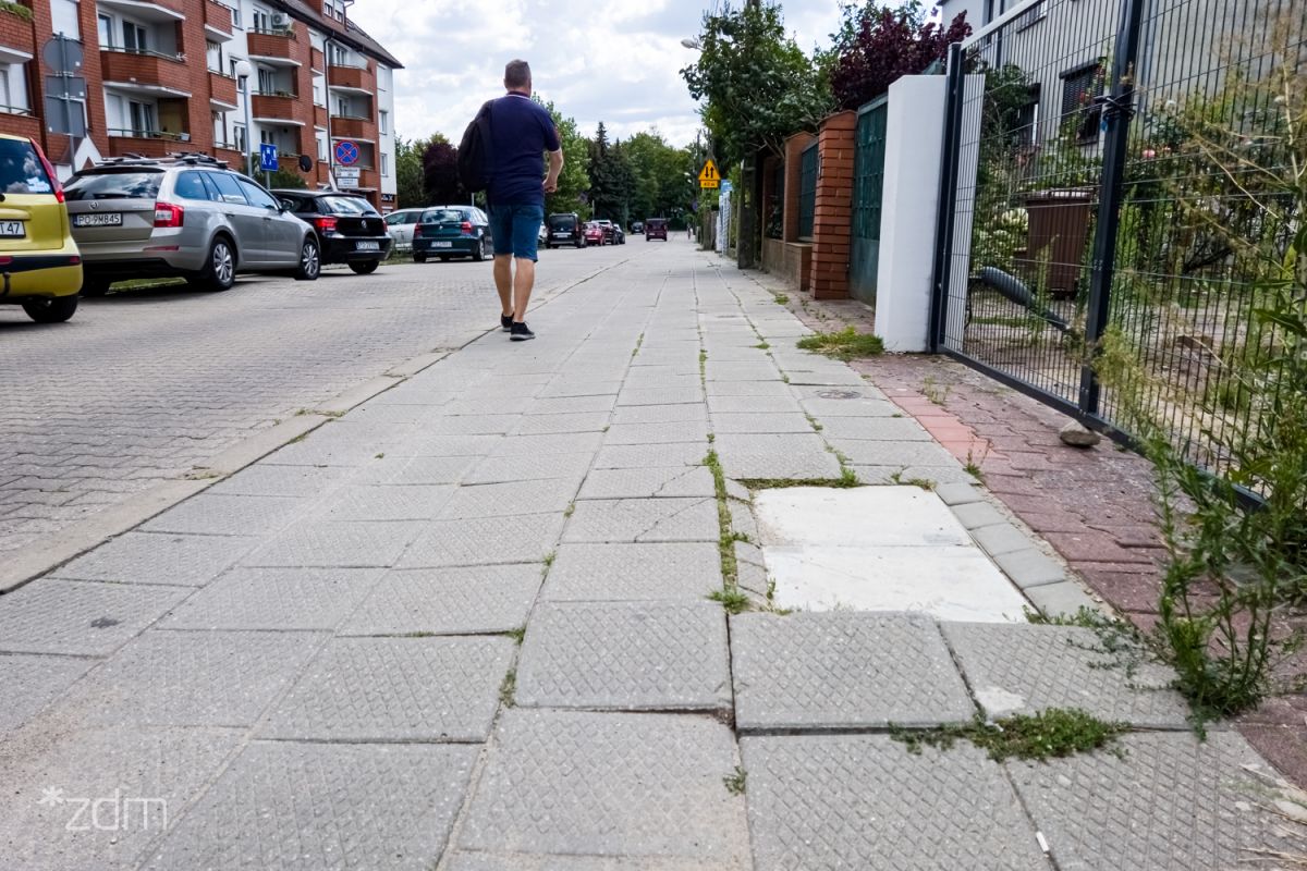 po lewej stronie zabudowania, następnie jezdnia. Centralnie widoczny chodnik z pieszym, po prawej fragment ogrodzenia.