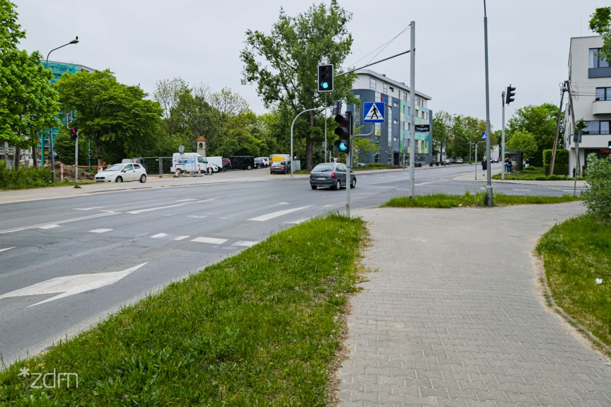 widok na ul. Piątkowską w kierunku północny, po lewej widać jezdnię, po prawej zieleń, znaki drogowe oraz fragment chodnika; w oddali skrzyżowanie z sygnalizacją świetlną oraz budynek.