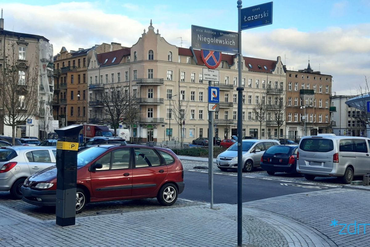 Okolice rynku Łazarskiego. Na pierwszym planie tabliczki z nazwami ulic na słupie. Za nimi parkujące samochody i zasłnięty folią parkomat