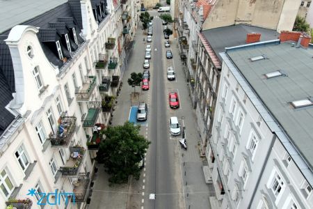 Zdjęcie z drona. Po prawej i lewej stronie kamienice, między nimi ulica z kilkoma drzeami i zaparkowanymi na jezdni samochodami 