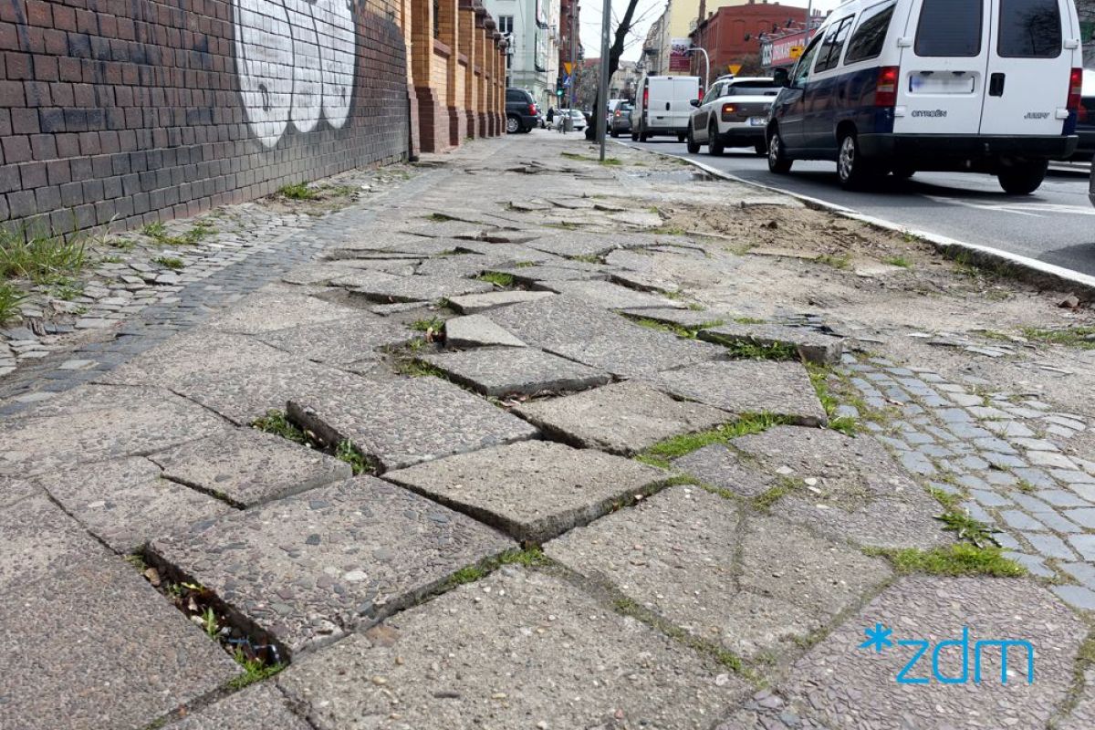 Zniszczony chodnik na ul. Wyspiańskiego, popękane i powykrzywioane płyty, w tle kamienice