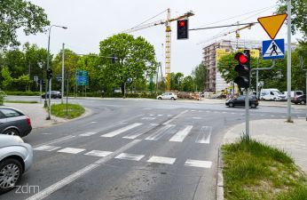 widok na skrzyżowanie, po prawej stronie fragment pojazdów stojących na czerwonym świetle, następnie przejście dla pieszych przez ul. Trójpole oraz sygnalizatory świetlne, następnie widać poprzecznie ulicę Piątkowską i pojazd; w oddali widać budowę, dźwig i powstające budynki.