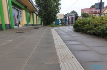 Chodnik z płyt betonowych z fakturą naprowadzającą na przystanek autobusowy. Na drugim planie zieleń i sklep.