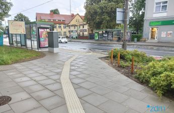 Chodnik z płyt betonowych z fakturą naprowadzającą na przystanek autobusowy. Na drugim planie przystanek i jezdnia.