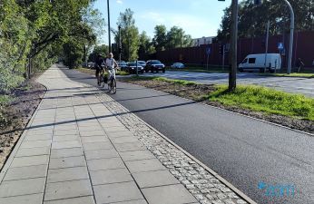 Chodnik z płyt betonowych i droga dla rowerów z asfaltu. W tle jadący rowerzyści.