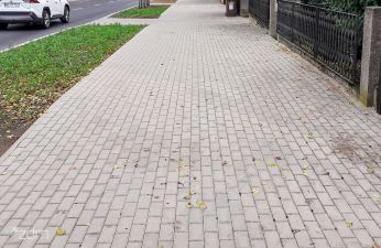Chodnik z kostki betonowej, po prawej stronie zieleni i płot, a po lewej pas zieleni i jezdnia.