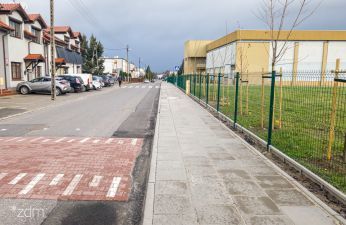 po lewej stronie jezdnia z progiem zwalniajacym po prawej stronie chodnik, ogrodzenie i fragment szkoły
