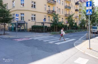 po lewej zabudowania, centralnie przejście dla pieszych przez ulicę przez które idzie kobieta, po prawej znak drogowy