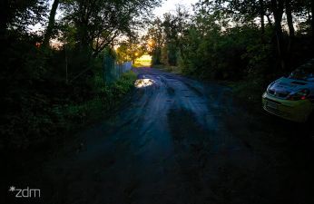 zdjęcie wykonane po ciemku, po lewej drzewa, centralnie ulica bez oświetlenia, po prawej fragment pojazdu