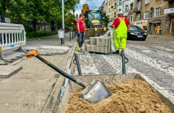 Widok na teren budowy, w tle taczka z piaskiem oraz osoby pracujące na przejściu dla pieszych
