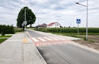 Ulica Garaszewo z przejściem dla pieszych i chodnikiem, w tle pola uprawne, drzewa i dom po prawej stronie