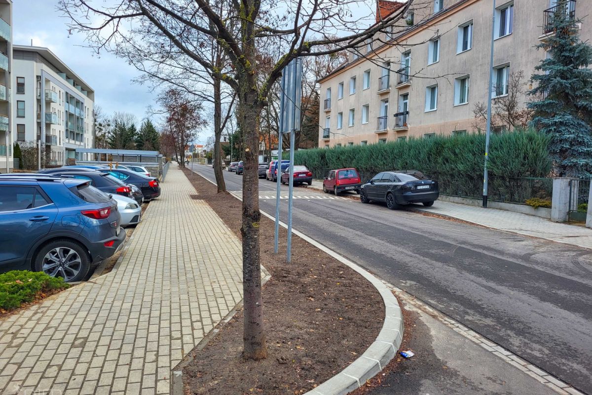 Ulica po przebudowie. Po lewej stronie zaparkowane samochody, a za nimi bloki. Następnie chodnik i pas gdzie będzie rosła zieleń. Na prawo jezdnia oraz bloki mieszkalne