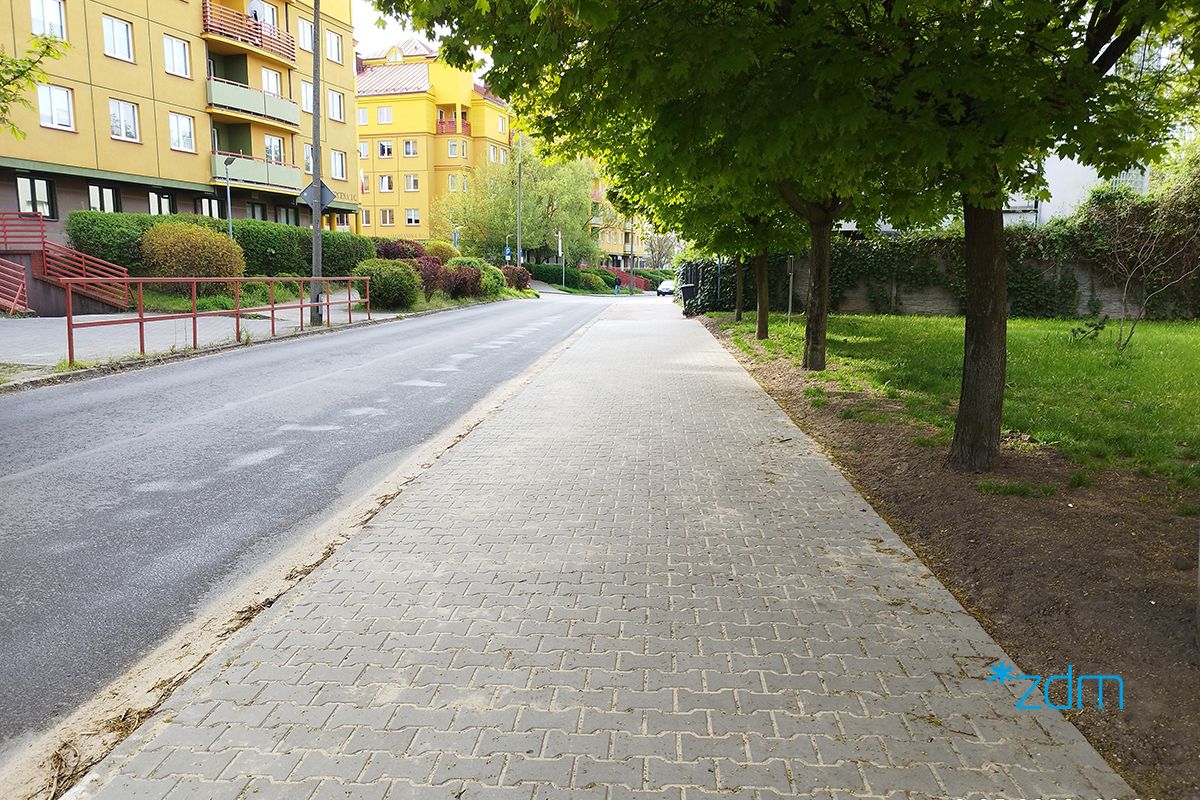 Widok chodnika z kostki betonowej na ul. Rylejewa w perspektywie zbieżnej, na drugim planie po lewej stronie blok mieszkalny, po prawej drzewa i teren zieleni