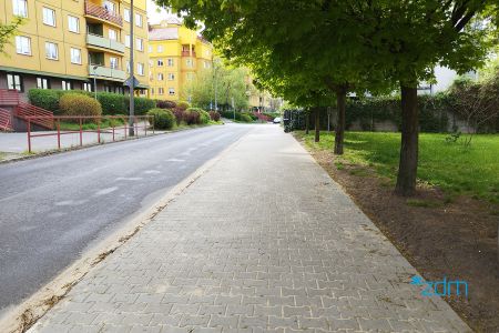 Widok chodnika z kostki betonowej na ul. Rylejewa w perspektywie zbieżnej, na drugim planie po lewej stronie blok mieszkalny, po prawej drzewa i teren zieleni