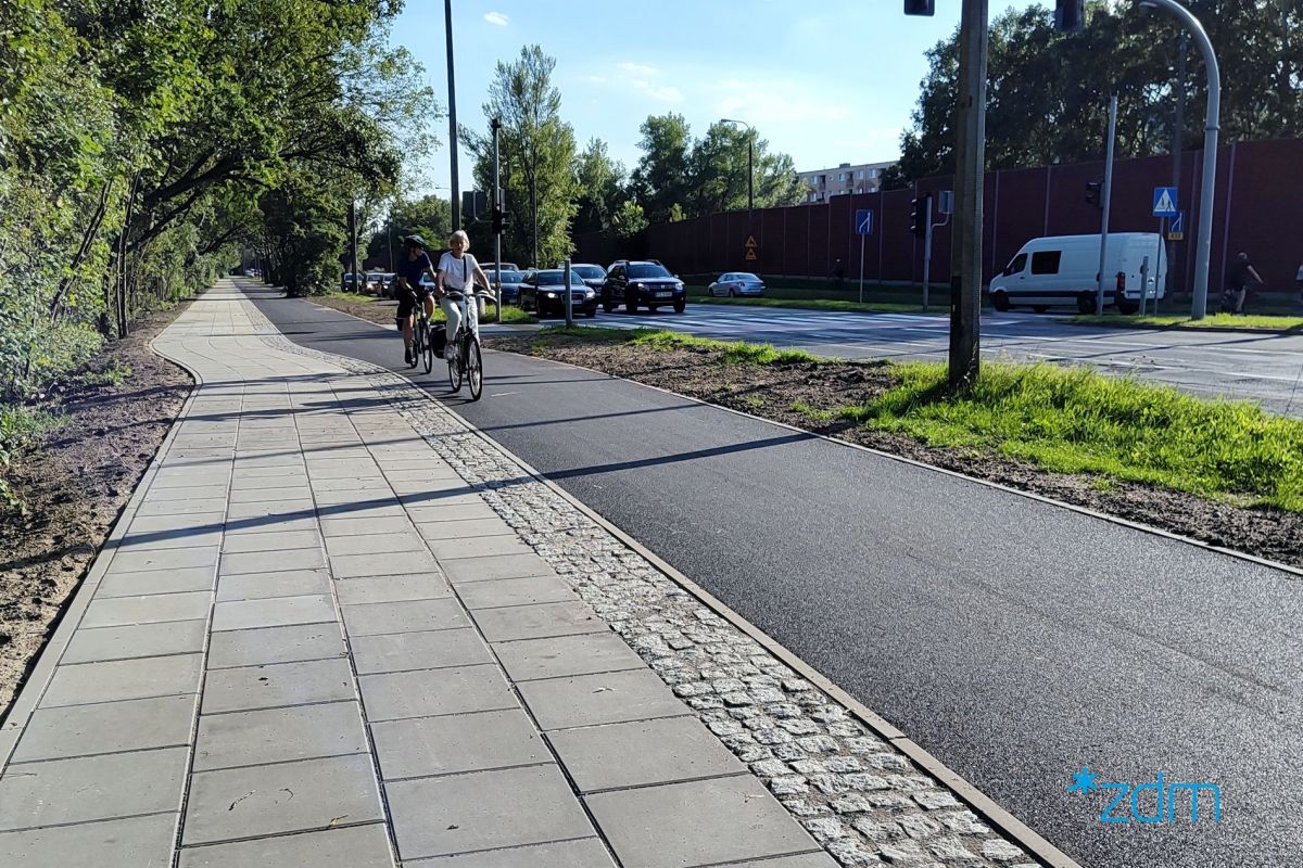 Chodnik z płyt betonowych i droga dla rowerów z asfaltu. W tle jadący rowerzyści.