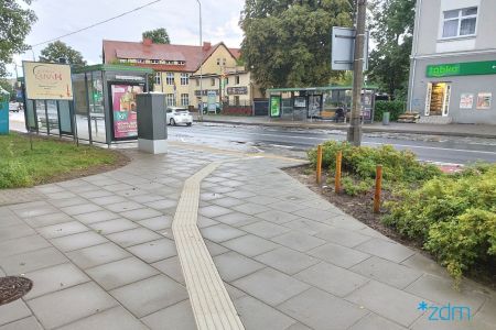 Chodnik z płyt betonowych z fakturą naprowadzającą na przystanek autobusowy. Na drugim planie przystanek i jezdnia.