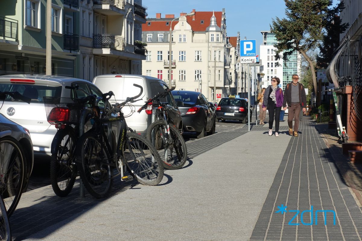 Wyremontowany chodnik na ul. Mickiewicza. Po lewej stronie zaparkowane samochody oraz rowery przyczepione do stojaków. Po środku chodnik na którym idzie para pieszych. Po prawej żywopłot