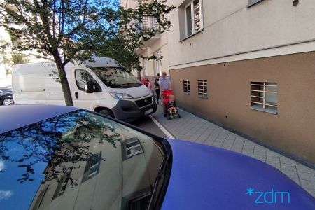 Po lewej stronie parkujące samochody, następnie chodnik z pieszymi i ściana budynku