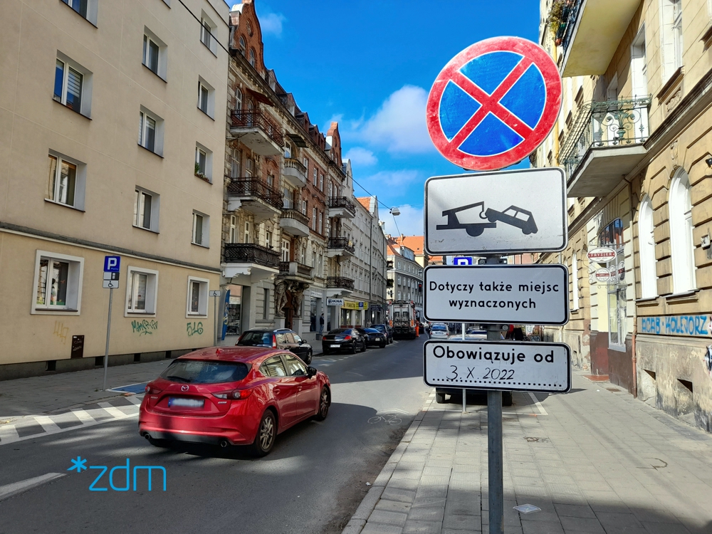 Znak zakaz zatrzymywania się ustawiony na chodniku. Za nim parkujące samochody