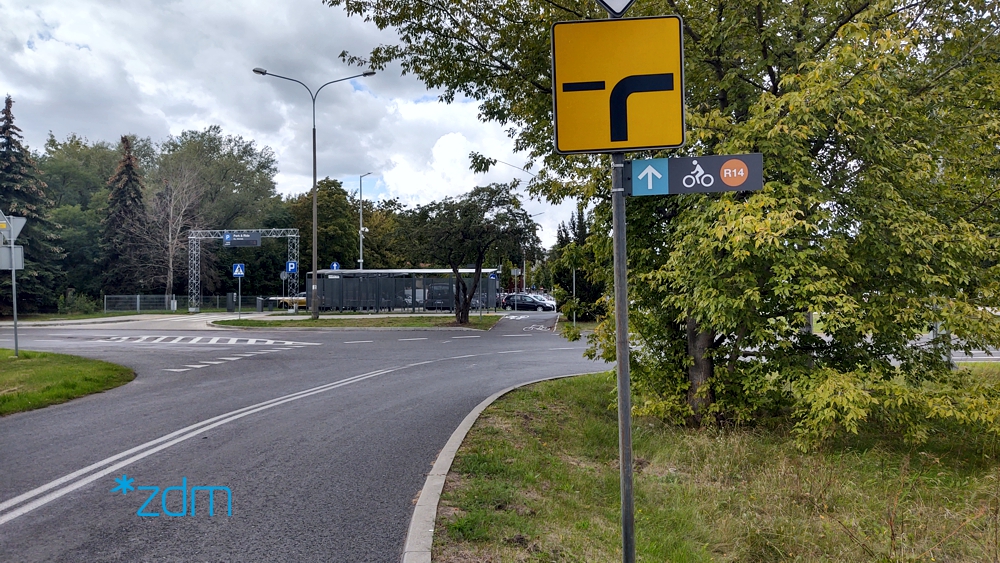 Znak drogowy dla samochodów oraz dla rowerow przed skrzyzowaniem. W tle parking oraz drzewa
